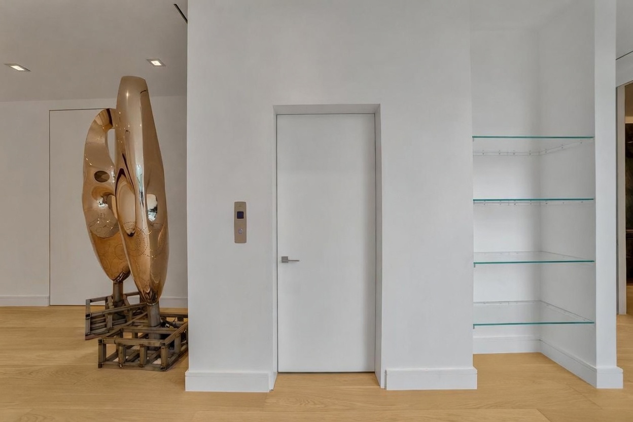 This interior view features a minimalist hallway with light wood flooring and crisp white walls. A central white door with a modern silver handle is flanked by a sculptural, metallic art piece on the left and a set of built-in glass shelving on the right. The space is illuminated by recessed ceiling lights, creating a clean, contemporary, and gallery-like atmosphere.