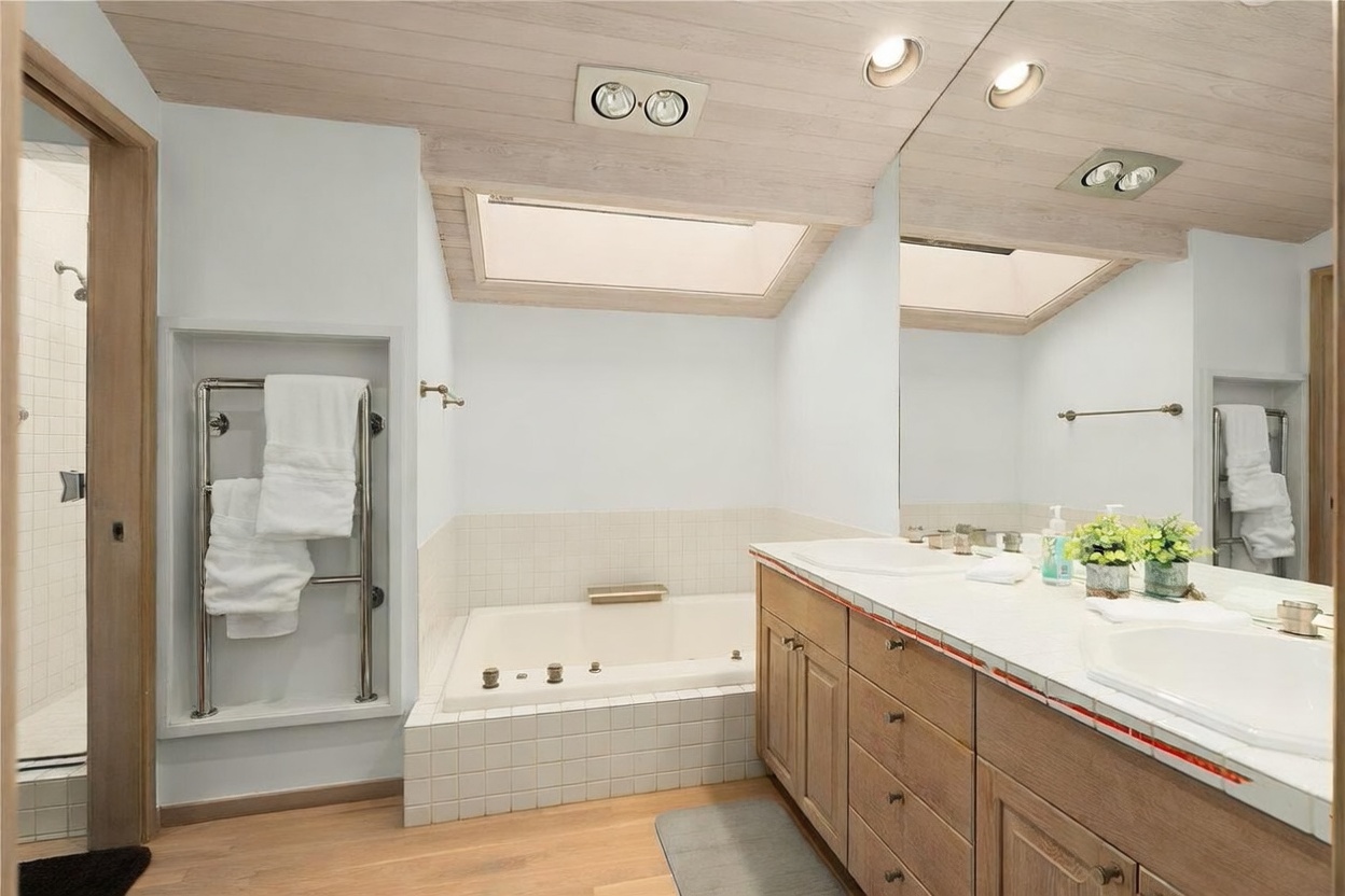 This primary bathroom features a light-toned wood vanity with dual sinks and a large mirror that reflects the space. A built-in soaking tub is positioned beneath a skylight, complemented by a separate walk-in shower area and a wall-mounted towel warmer. The room is finished with light-colored tile surfaces and a vaulted wood-paneled ceiling, creating a bright and airy atmosphere.