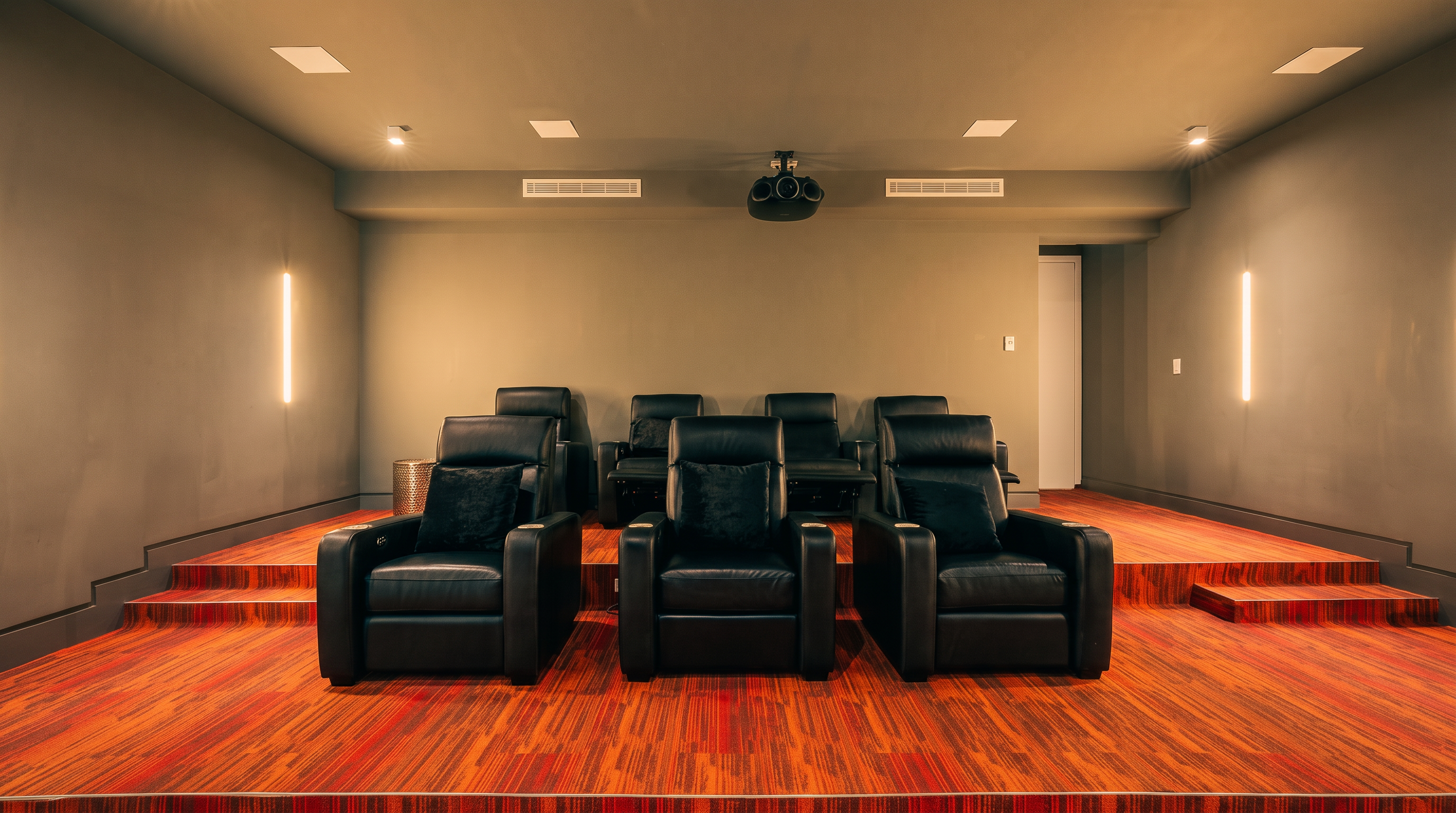 This dedicated home theater room features a tiered seating arrangement with six black leather recliners, three in the front row and three in the back. The room is designed with a modern aesthetic, showcasing a bold, multi-toned orange and brown patterned carpet and neutral gray walls. A projector is mounted to the ceiling, and vertical light fixtures on the side walls provide ambient illumination for a cinematic experience.