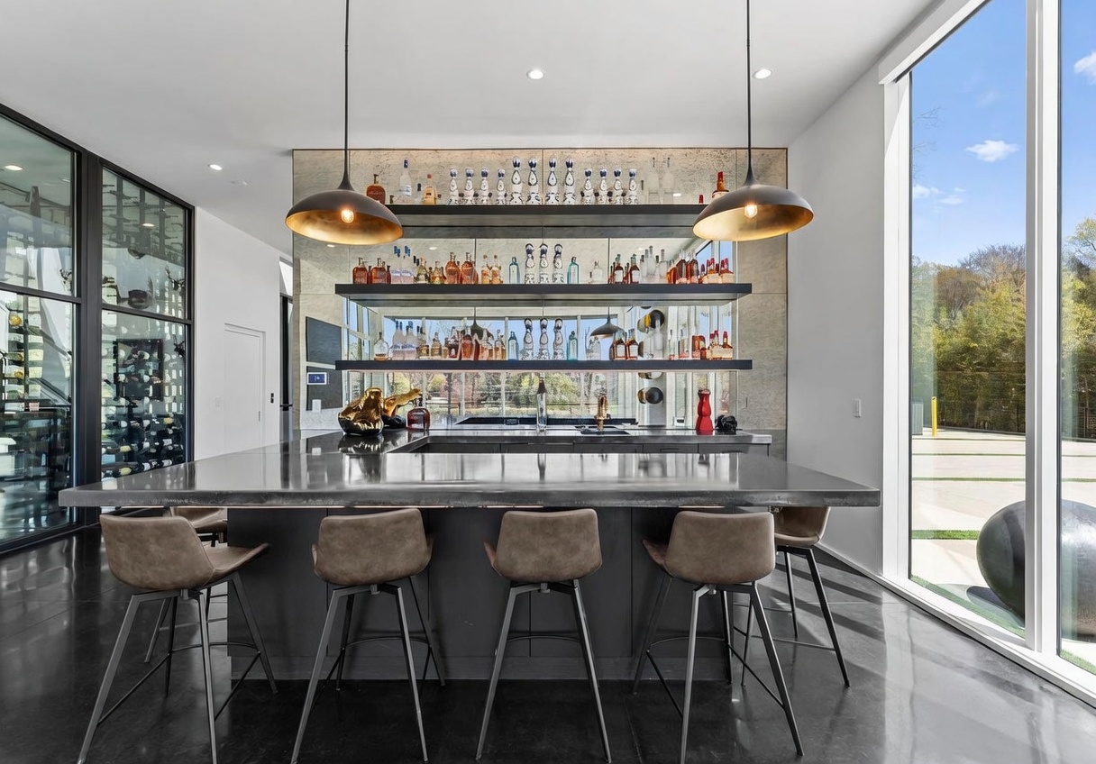 This sophisticated home bar area features a large, dark-toned island with bar seating and a mirrored back wall lined with illuminated floating shelves displaying a collection of spirits. The space is anchored by two large, metallic pendant lights and is flanked by a glass-enclosed wine storage unit on the left and expansive glass walls on the right that provide a view of the outdoors. The sleek, modern aesthetic is enhanced by polished dark flooring and a clean, minimalist design approach.