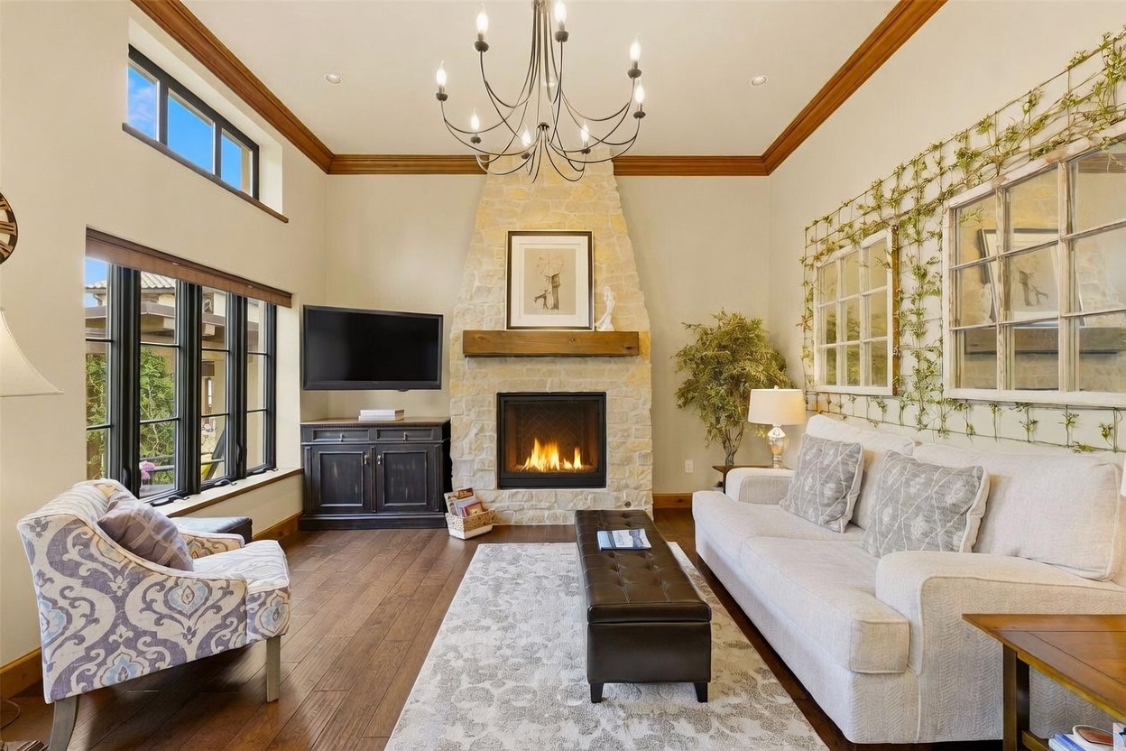 This inviting living room features a prominent stone-clad chimney breast with a wooden mantel as its central focal point. The space is furnished with a light-colored sofa, a patterned armchair, and a leather ottoman, all arranged on a textured area rug over dark wood flooring. Large windows and a transom window allow natural light to fill the room, which is accented by a decorative wall trellis and a classic chandelier.