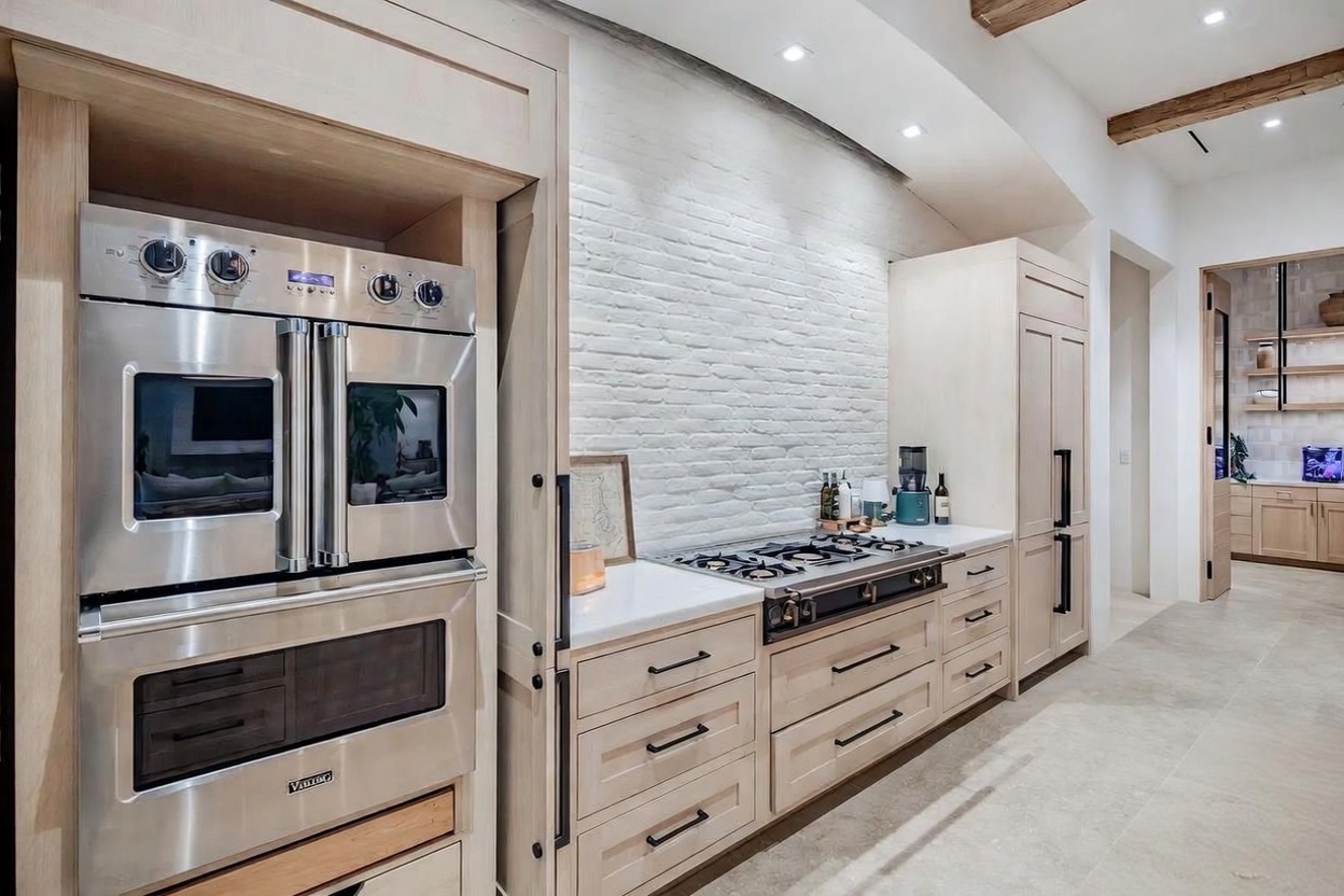 This bright, modern kitchen features light wood cabinetry, a white brick backsplash, and high-end stainless steel appliances, including a Viking double oven. The space is accented by exposed wooden ceiling beams and a neutral stone floor, creating a warm and sophisticated atmosphere. The perspective captures the main cooking area, showcasing the seamless integration of professional-grade appliances with custom cabinetry.