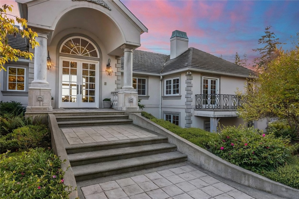 This elegant home features a grand entryway with a prominent arched portico supported by classic columns, leading to a set of double glass-paned doors. A wide, tiered concrete staircase with integrated lighting leads up to the entrance, flanked by lush landscaping and a small balcony with decorative iron railings on the right. The exterior is finished in a neutral tone with stone accents, set against a vibrant, colorful sunset sky.