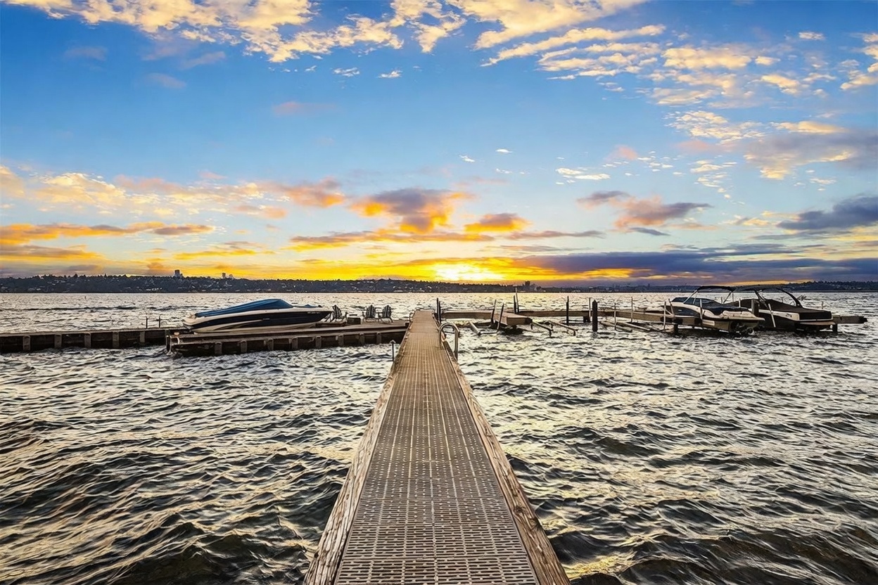This scenic waterfront view captures a long, metal-grated pier extending into a calm lake at sunset. The horizon is illuminated by a vibrant, golden-hued sky with scattered clouds, while several boats are moored along the sides of the dock. The perspective is centered and low, drawing the viewer's eye directly toward the tranquil water and the distant shoreline.