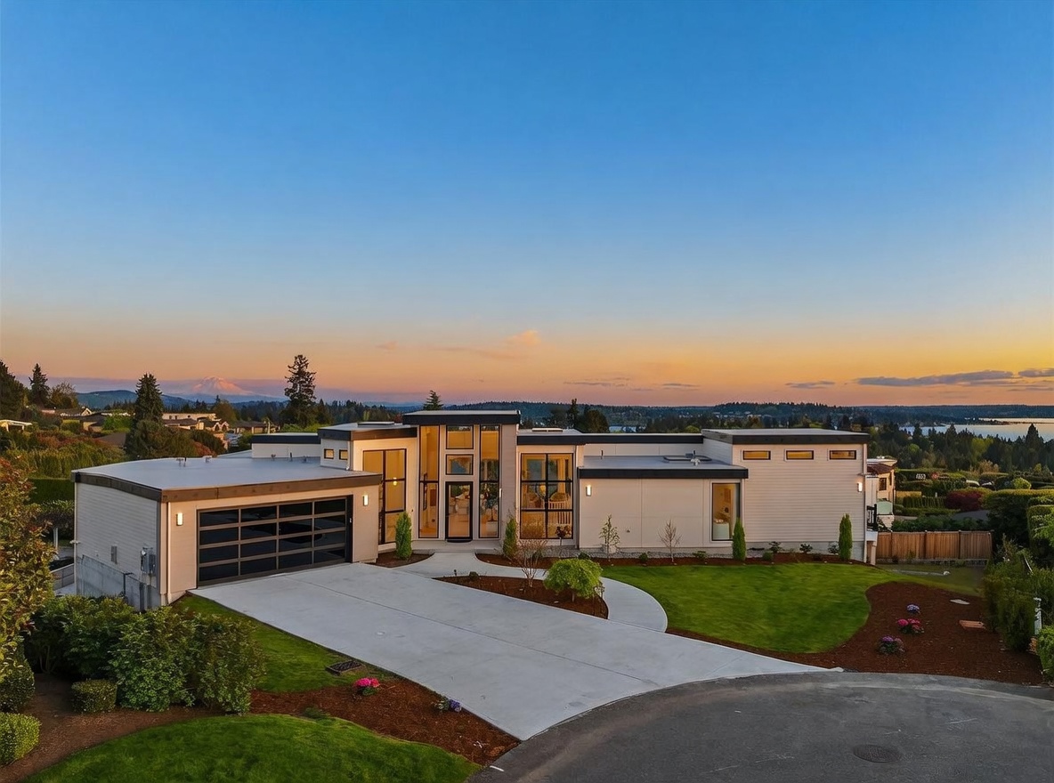 This modern, single-story residence features a striking architectural design with clean lines, a flat roof, and a prominent central entryway framed by large windows. The exterior is finished in light-toned horizontal siding, complemented by a multi-panel glass garage door and a concrete driveway that curves toward the front entrance. Set against a scenic sunset backdrop with a distant mountain peak, the property includes a well-manicured lawn and professional landscaping, creating a sophisticated and inviting curb appeal.