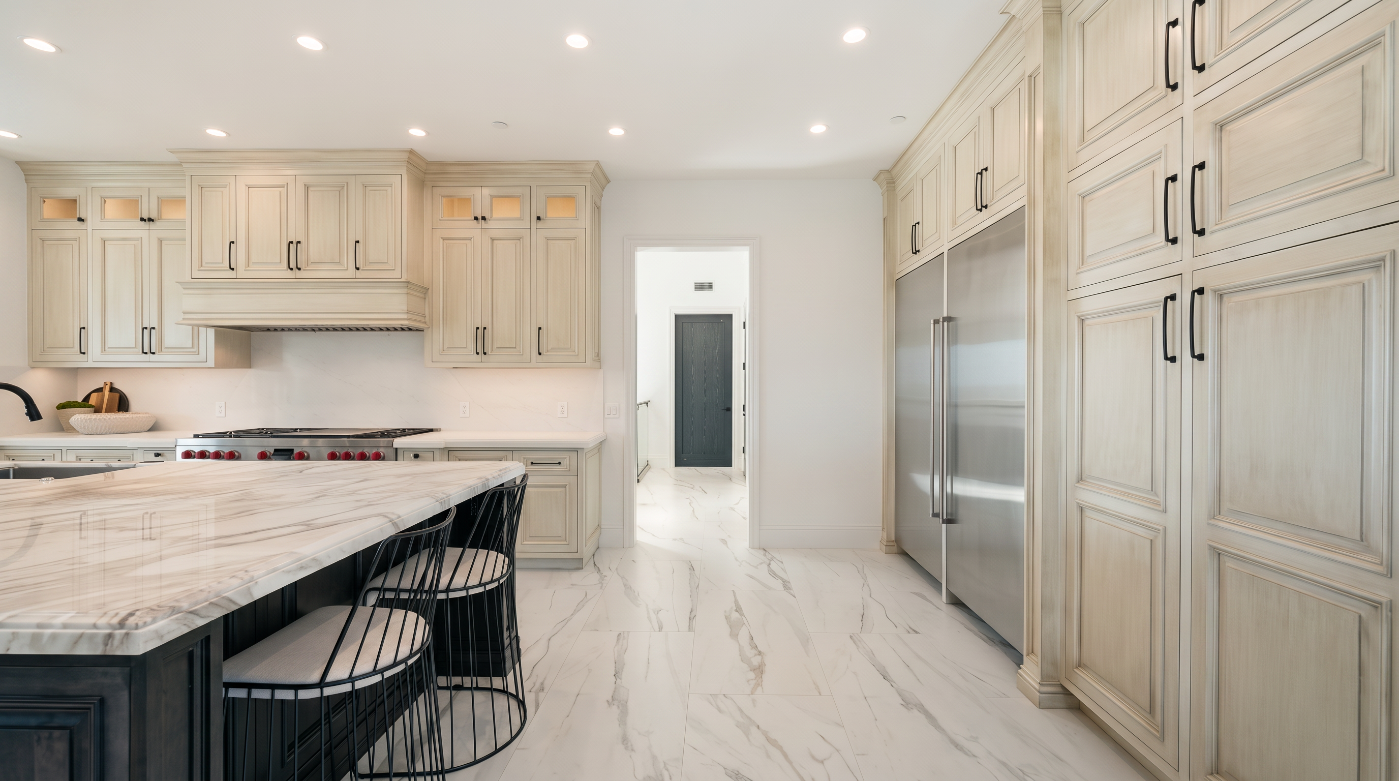 This bright, modern kitchen features a large central island with a patterned stone countertop and dark base cabinetry, complemented by light-toned custom cabinetry throughout. A professional-style range is set against a tiled backsplash, while a large stainless steel refrigerator is integrated into the cabinetry wall. The space is finished with light-colored stone flooring and recessed lighting, creating an open and airy atmosphere that leads toward a doorway in the background.