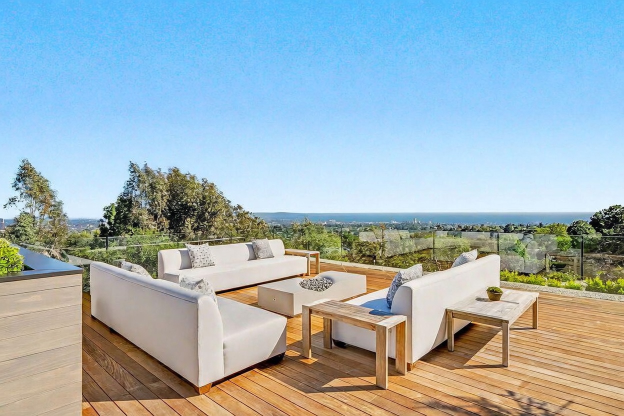 This expansive wooden deck offers a sophisticated outdoor living space with a panoramic view of the coastline and horizon. The area is furnished with two modern white sofas arranged around a central low-profile coffee table, complemented by light wood side tables. A glass railing ensures an unobstructed view, creating a seamless transition between the deck and the scenic landscape beyond.