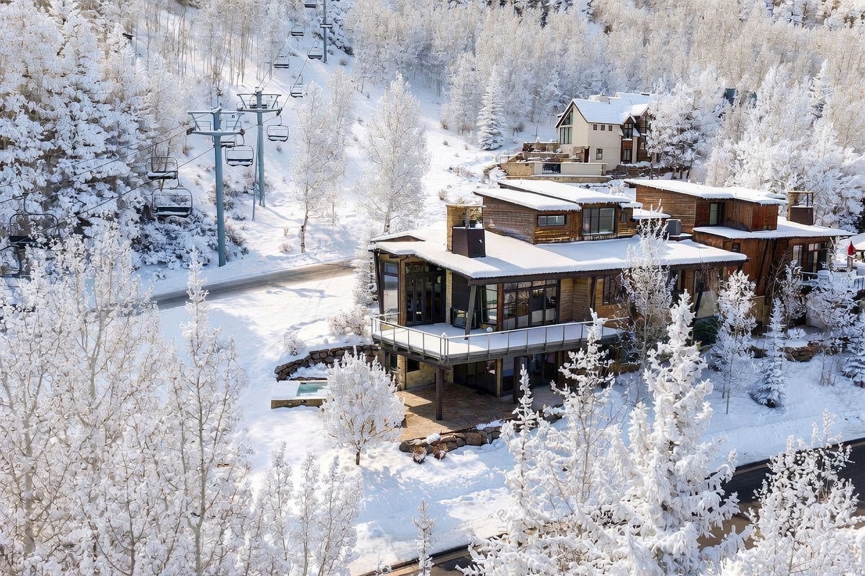 This aerial perspective captures a modern mountain residence nestled within a snow-covered, forested landscape. The home features a multi-level design with wood siding, expansive glass windows, and a spacious deck overlooking the snowy terrain. A ski lift runs directly adjacent to the property, emphasizing its prime location for winter sports enthusiasts.
