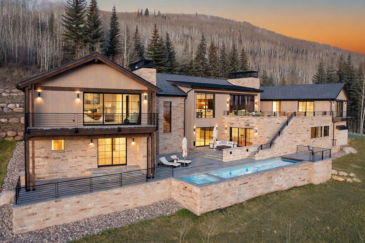 This expansive mountain estate features a sophisticated blend of natural stone and vertical wood siding, set against a backdrop of dense evergreen and aspen forests. The rear exterior showcases a multi-level terrace with a sleek, rectangular swimming pool and integrated spa, complemented by outdoor seating areas and a staircase leading to the upper deck. The architecture emphasizes a seamless connection between indoor and outdoor living through large windows and sliding glass doors, creating a luxurious and serene retreat.