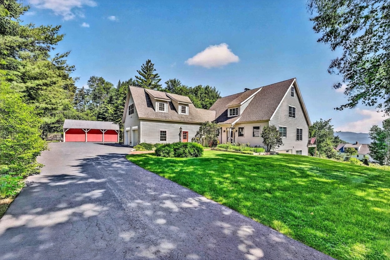 This image captures a charming, multi-level suburban home with light-colored siding and a steep, gabled roofline. A long, paved driveway leads toward a detached red-painted garage structure, while a lush green lawn stretches across the foreground. The scene is set under a bright blue sky with scattered clouds, highlighting the home's inviting curb appeal and scenic, tree-lined surroundings.