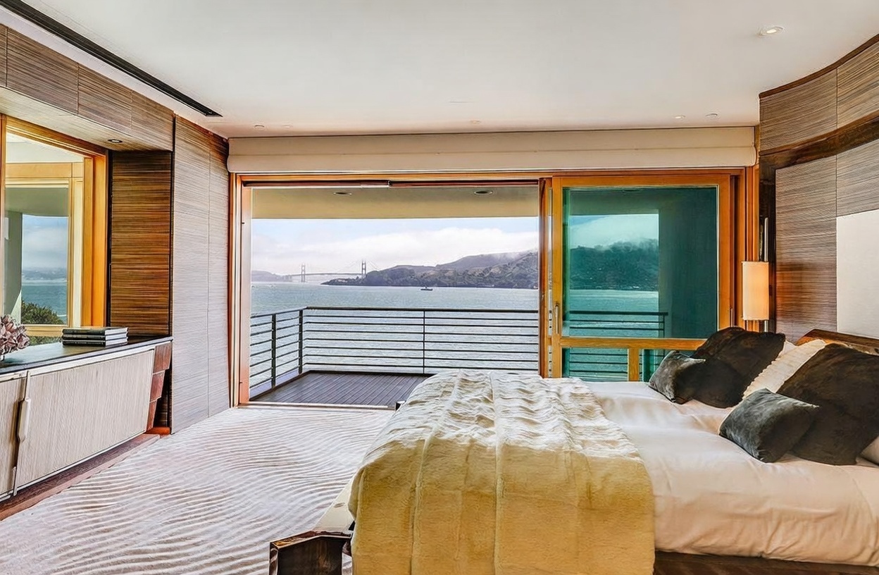 This luxurious primary bedroom features a minimalist aesthetic with wood-paneled walls and a plush bed dressed in neutral tones. A large sliding glass door opens onto a private balcony, framing a stunning, unobstructed view of the Golden Gate Bridge and the bay. The room is bathed in natural light, creating a serene and sophisticated atmosphere that emphasizes the connection between the interior space and the iconic waterfront scenery.