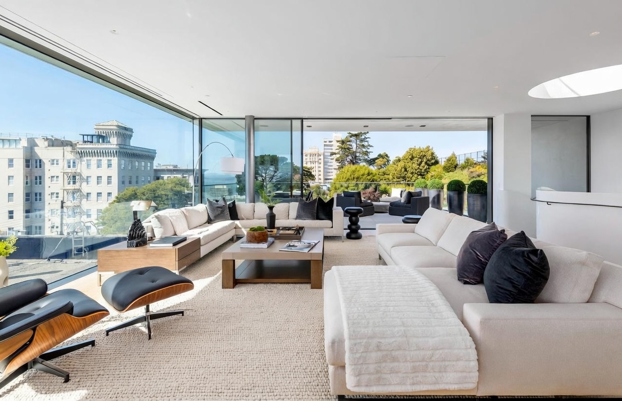 This expansive, modern living room features a bright, open-concept layout defined by floor-to-ceiling glass walls that offer panoramic city and greenery views. The space is furnished with two large, neutral-toned sectional sofas arranged around a central wooden coffee table, complemented by a classic lounge chair and ottoman. The room flows seamlessly onto an outdoor terrace, creating a sophisticated indoor-outdoor living experience bathed in natural light.