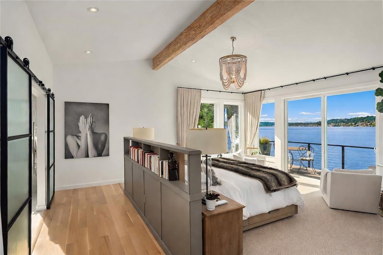 This primary bedroom features a sophisticated open-concept layout with a central partition wall that doubles as a bookshelf, separating the sleeping area from the entryway. The room is highlighted by a vaulted ceiling with a prominent exposed wood beam, a beaded chandelier, and large sliding glass doors that open to a private balcony overlooking the water. The space is finished with light wood flooring and neutral tones, creating a serene and luxurious atmosphere.