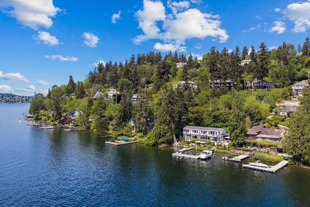 This aerial perspective captures a serene waterfront property nestled along a lush, tree-covered hillside. The scene features a multi-level residence with expansive decks overlooking the water, complemented by private docks and boat slips extending into the lake. The surrounding landscape is densely wooded, creating a private and tranquil atmosphere typical of a premium lakeside retreat.