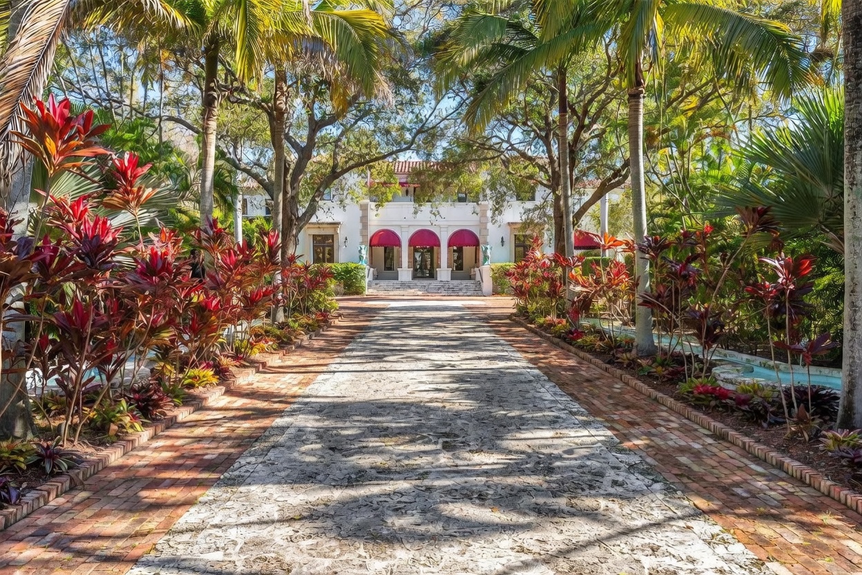 This grand estate features a long, stone-paved driveway lined with lush tropical landscaping and mature trees, leading directly to the main entrance. The white stucco facade is accented by three prominent red arched awnings over the entry doors, creating a striking Mediterranean-inspired aesthetic. The perspective is centered and symmetrical, emphasizing the impressive scale and private, park-like setting of the property.