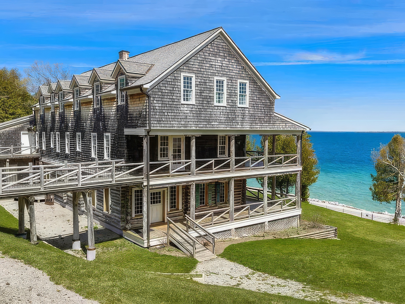 This expansive, multi-story shingled home features a classic coastal aesthetic with wrap-around porches on two levels and a raised wooden walkway connecting to an adjacent structure. The exterior is clad in weathered wood shingles, complemented by numerous white-trimmed windows and a gabled roofline. Set against a vibrant blue sky and overlooking a clear, turquoise-toned body of water, the property offers a serene and picturesque waterfront setting.