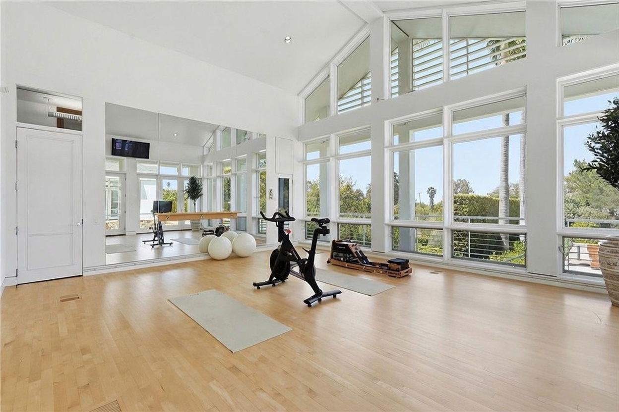 This bright and airy home gym features light wood flooring and a wall of mirrors that enhances the sense of space. Large windows span the wall, providing abundant natural light and views of the surrounding greenery. The room is equipped with a stationary bike, a rowing machine, and exercise balls, creating a functional and inviting workout environment.