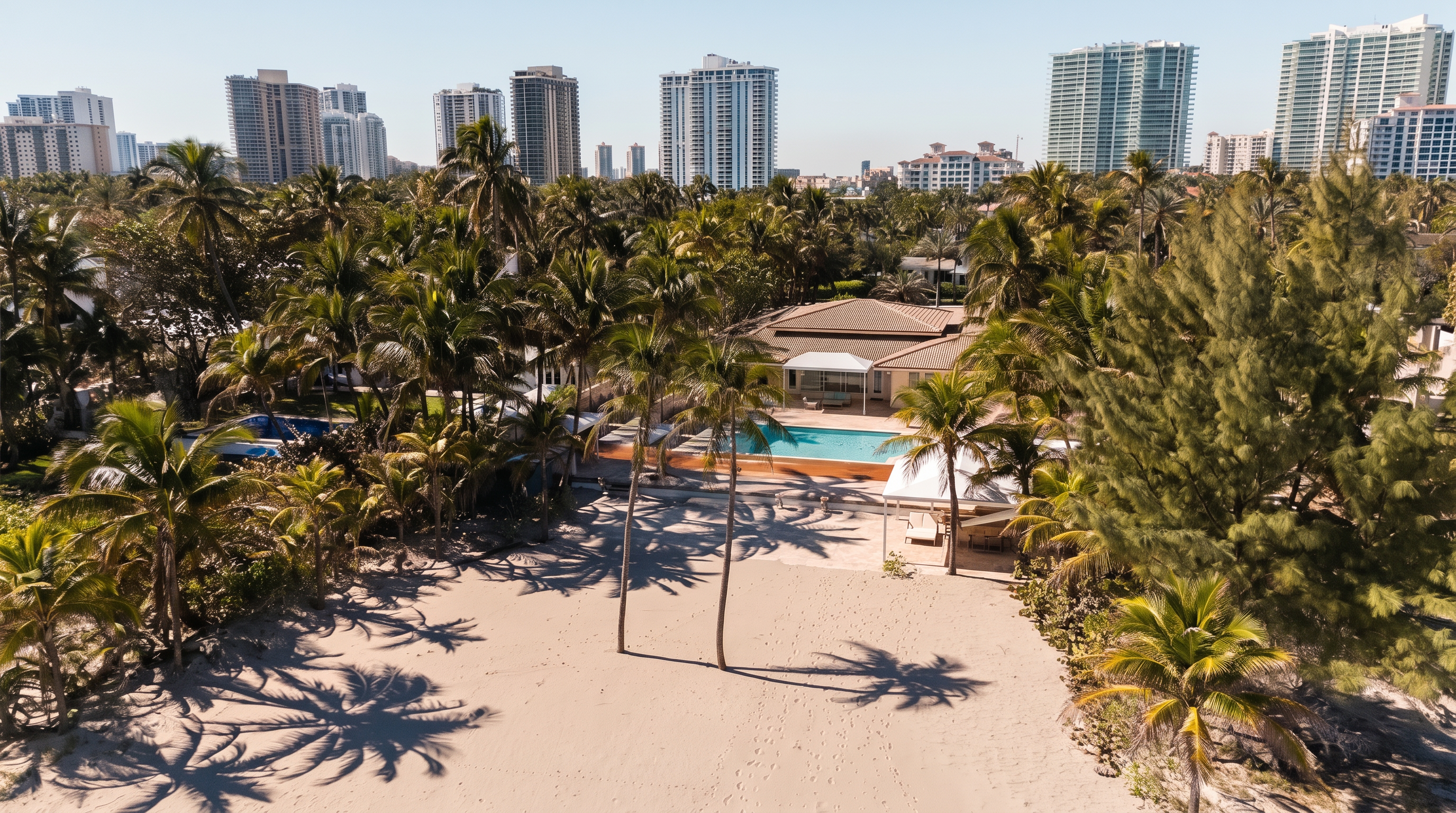 This aerial perspective captures a beachfront property featuring a private swimming pool and a spacious patio area nestled among lush palm trees. The home is situated directly on the sand, offering a seamless transition from the residence to the beach, with a backdrop of high-rise urban buildings. The scene conveys a luxurious, tropical lifestyle with a clear view of the property's prime coastal location.