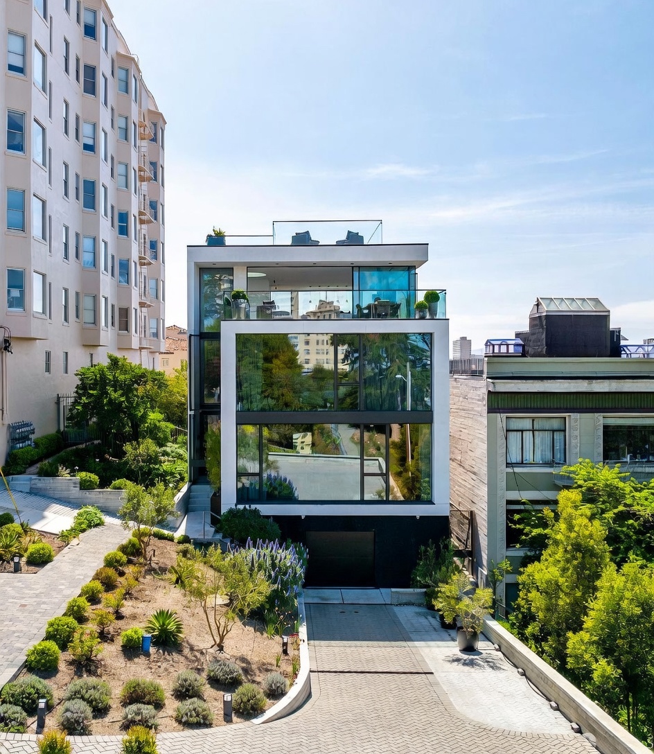 This modern, multi-level residence features a striking contemporary design with extensive glass facades and clean, geometric lines. The property includes a private driveway leading to a garage, a landscaped front garden with drought-tolerant plantings, and multiple levels of outdoor terrace space. The structure is nestled between a taller apartment building and a more traditional neighboring home, creating a unique contrast in architectural styles.
