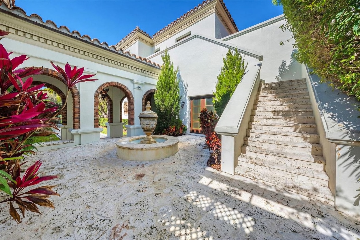 This Mediterranean-style courtyard features a stone-paved patio centered around a classic tiered fountain. A grand stone staircase with a solid balustrade leads to an upper level, while arched brick-lined openings provide architectural interest and access to the surrounding grounds. Lush greenery and vibrant red-leafed plants frame the space, creating a serene and elegant outdoor retreat.