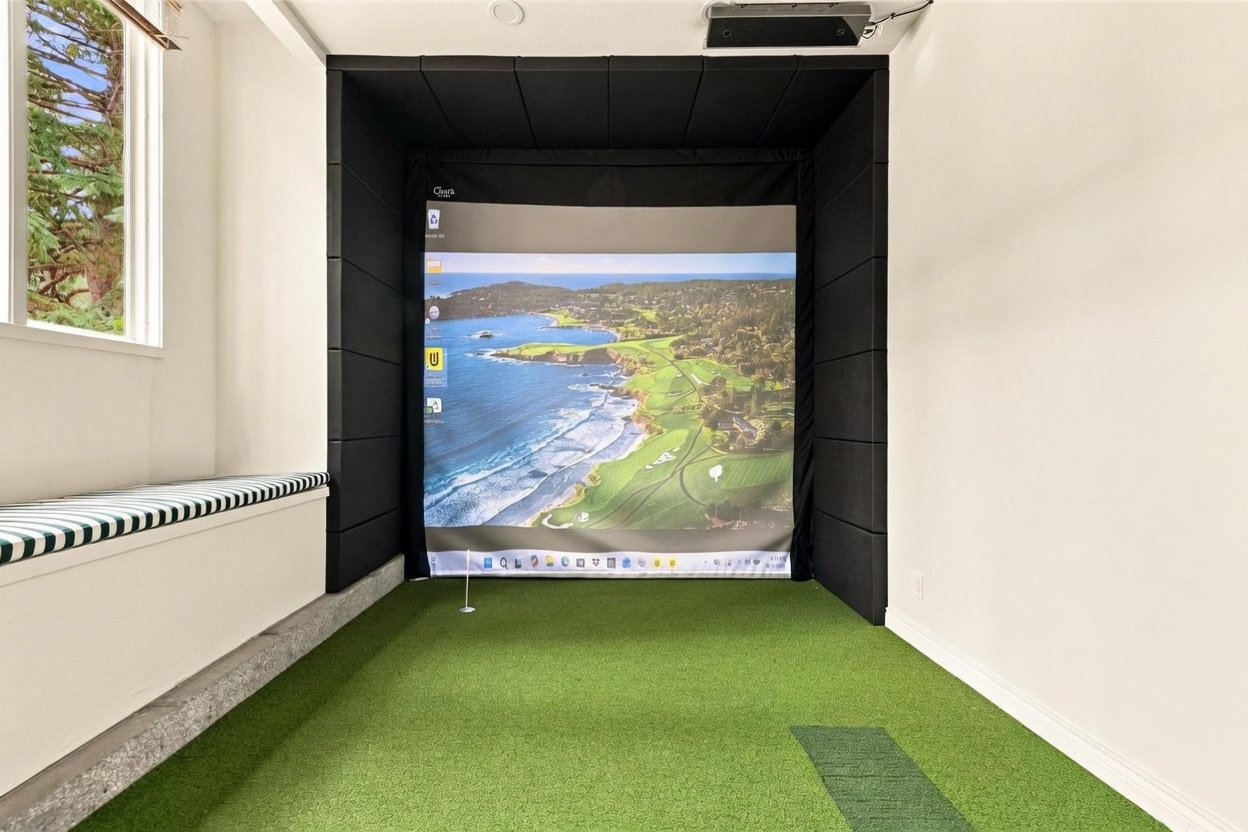 This image showcases a dedicated indoor golf simulator room featuring a large projection screen framed by black acoustic padding. The space is equipped with artificial turf flooring, a hitting mat, and a small putting hole, creating a realistic practice environment. To the left, a built-in bench with a striped cushion sits beneath a window, providing a comfortable seating area for spectators.