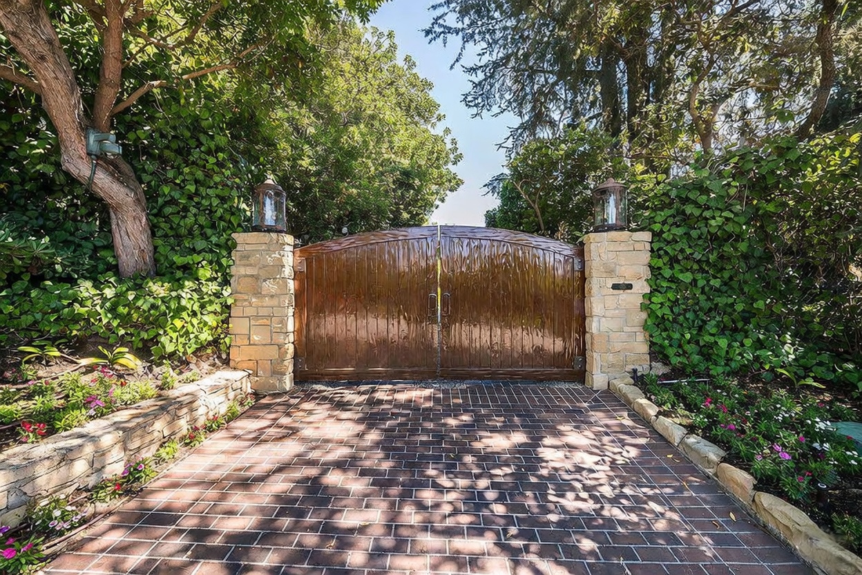 This image captures a private gated entrance featuring a pair of arched, textured wood doors set between two stone-clad pillars. The driveway is paved with dark, rectangular bricks, leading up to the gate, which is flanked by lush greenery and mature trees. The scene is framed by a stone retaining wall and vibrant landscaping, creating a secluded and secure aesthetic for the property.