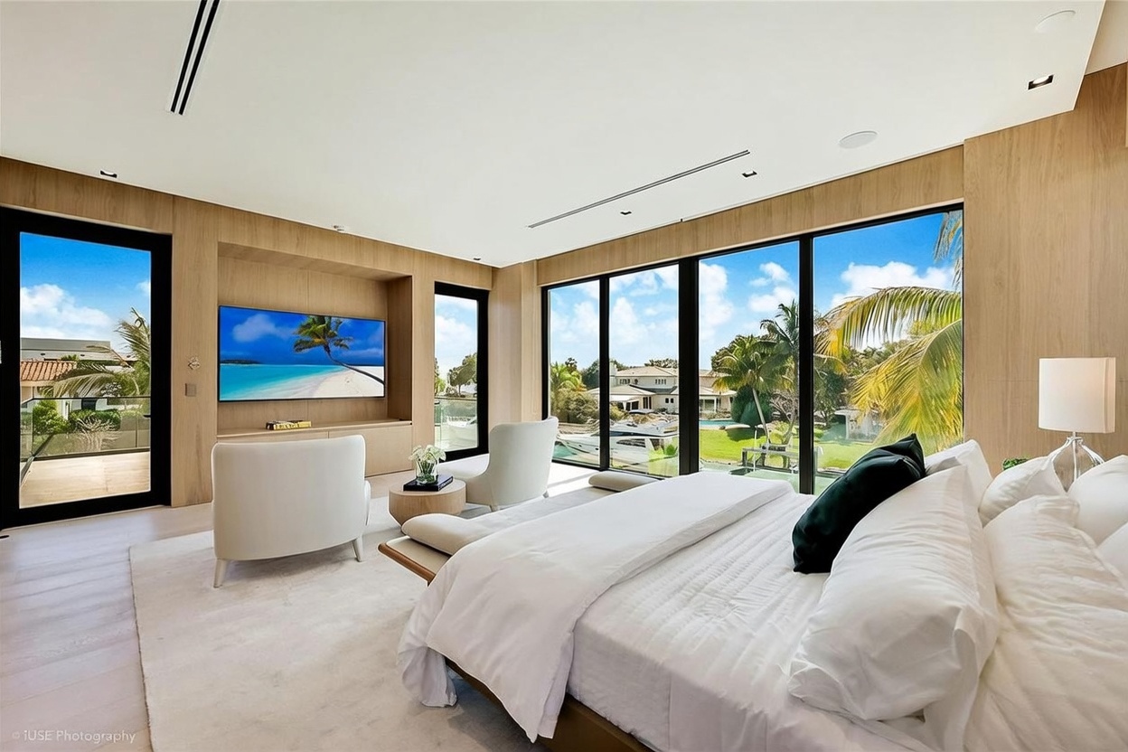 This luxurious primary bedroom features a minimalist aesthetic with light wood wall paneling and a neutral color palette. The room is anchored by a large bed facing a built-in media wall with a television, while floor-to-ceiling windows provide expansive views of the waterfront and lush landscaping outside. Two comfortable armchairs are positioned near the windows, creating a serene seating area that emphasizes the bright, airy atmosphere of the space.
