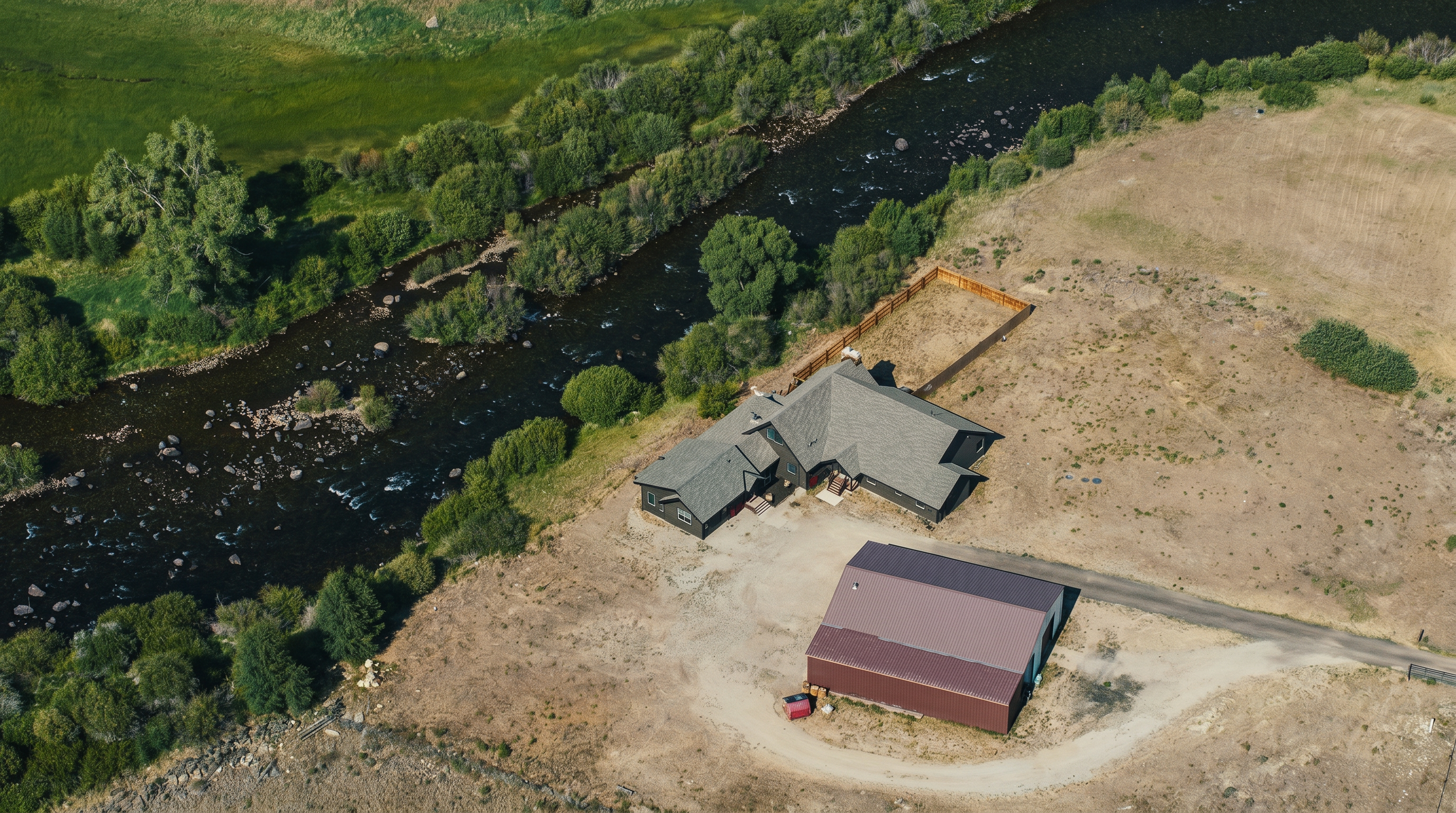 This high-angle aerial view captures a rural property situated alongside a winding river, featuring a multi-gabled house and a large detached metal-roofed barn. The landscape is characterized by expansive open fields, lush riparian vegetation along the water's edge, and a gravel driveway connecting the structures. The perspective emphasizes the secluded, natural setting and the generous acreage surrounding the home.