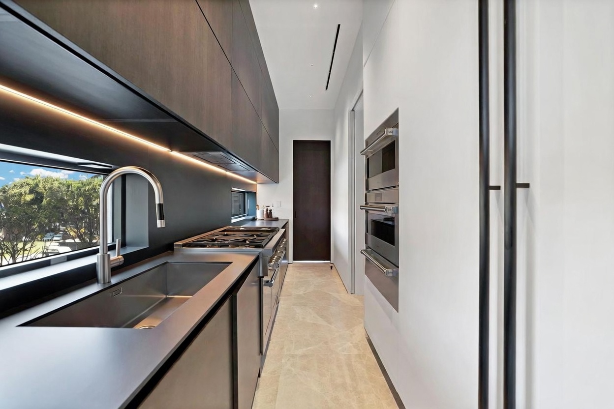 This sleek, modern galley-style kitchen features dark, minimalist cabinetry paired with a stainless steel sink and integrated appliances. A long, horizontal window above the counter provides natural light and a view of the outdoors, while under-cabinet lighting highlights the workspace. The perspective looks down the narrow corridor, showcasing the clean lines, stone flooring, and a tall, dark door at the far end.