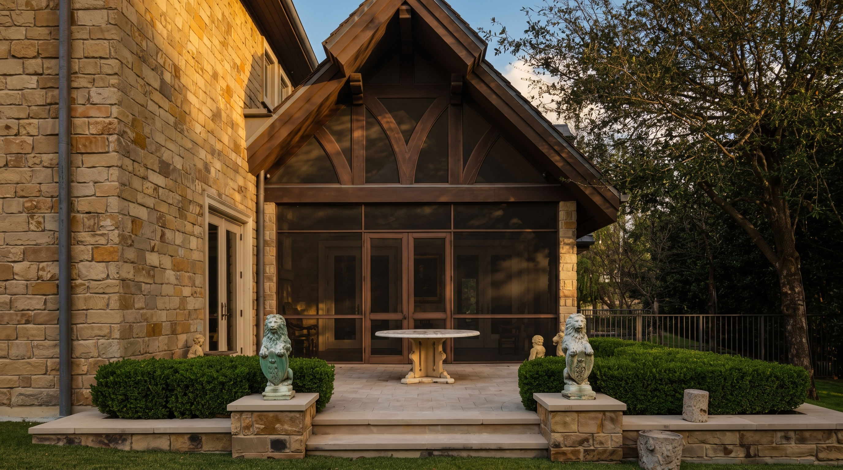 This image captures a serene outdoor patio space featuring a screened-in porch with a gabled roof and decorative timber framing. The patio is paved with light-toned stone and includes a central circular table, flanked by two stone lion statues atop low masonry walls that border manicured boxwood hedges. The scene is set against a backdrop of a stone-clad home exterior and mature trees, creating a sophisticated and private backyard atmosphere.
