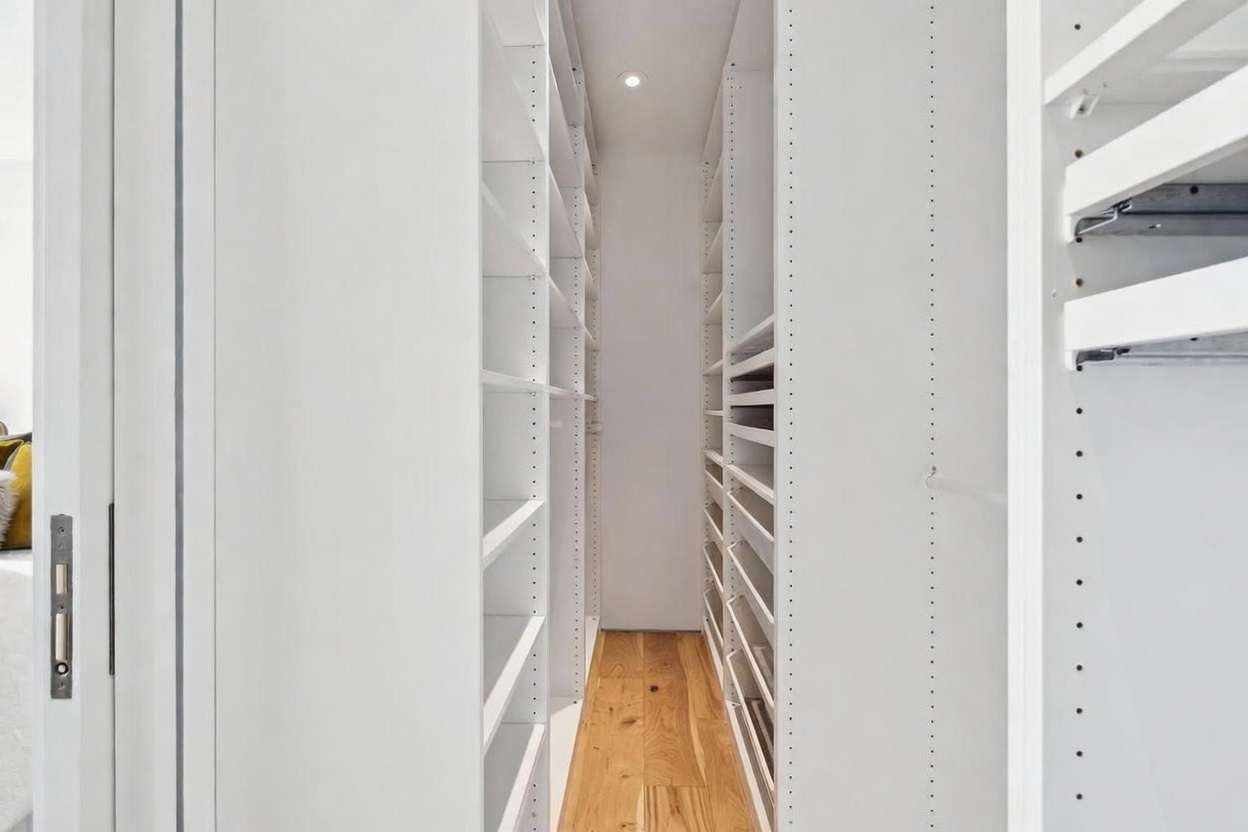 This walk-in closet features a clean, minimalist design with white shelving units lining both sides of a narrow, light wood-floored corridor. The space is illuminated by a single recessed ceiling light, highlighting the adjustable shelving tracks and the organized storage layout. The perspective is a straight-on view looking down the center of the closet, emphasizing its depth and efficient use of space.