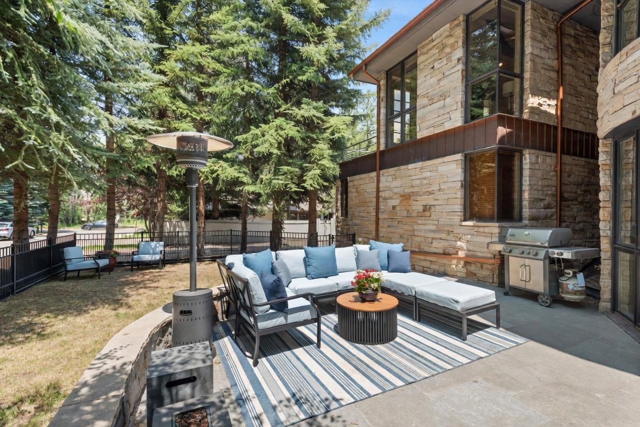 This inviting outdoor patio space features a stone-clad exterior wall and a comfortable seating area arranged on a striped area rug. The space is equipped with a stainless steel grill, a tall outdoor heater, and a low stone retaining wall that separates the patio from the grassy yard. Large windows on the multi-story stone home provide a view into the interior, creating a seamless transition between indoor and outdoor living.