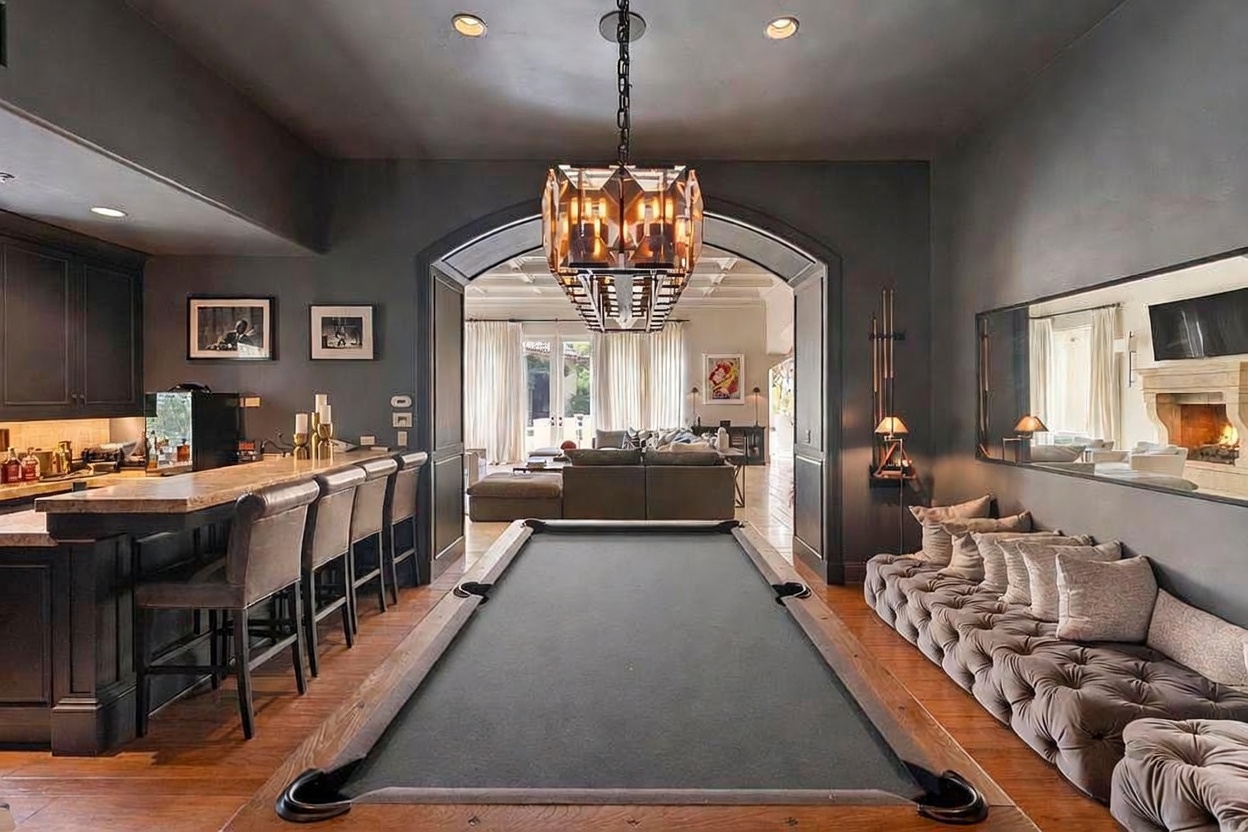 This interior shot showcases a luxurious living room with a pool table as a central feature. The room is decorated in dark, sophisticated tones, complemented by a unique chandelier and a comfortable-looking tufted bench along the wall. An arched doorway leads to another living space, creating an open and inviting atmosphere.