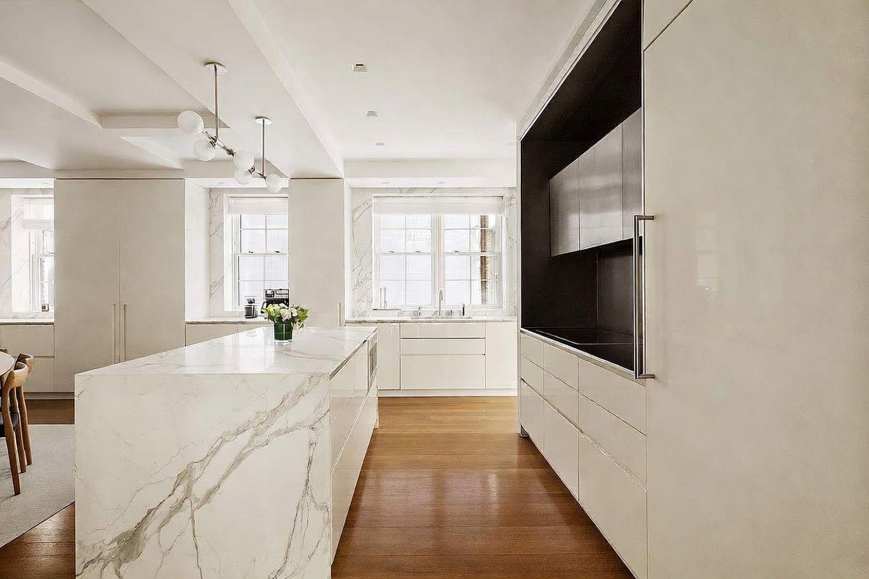 This sleek, contemporary kitchen features a large central island with a prominent stone waterfall edge and a light wood floor. The space is defined by minimalist white cabinetry and a striking dark-toned recessed wall section that houses integrated appliances. Large windows at the rear allow natural light to flood the room, creating an airy and sophisticated atmosphere.
