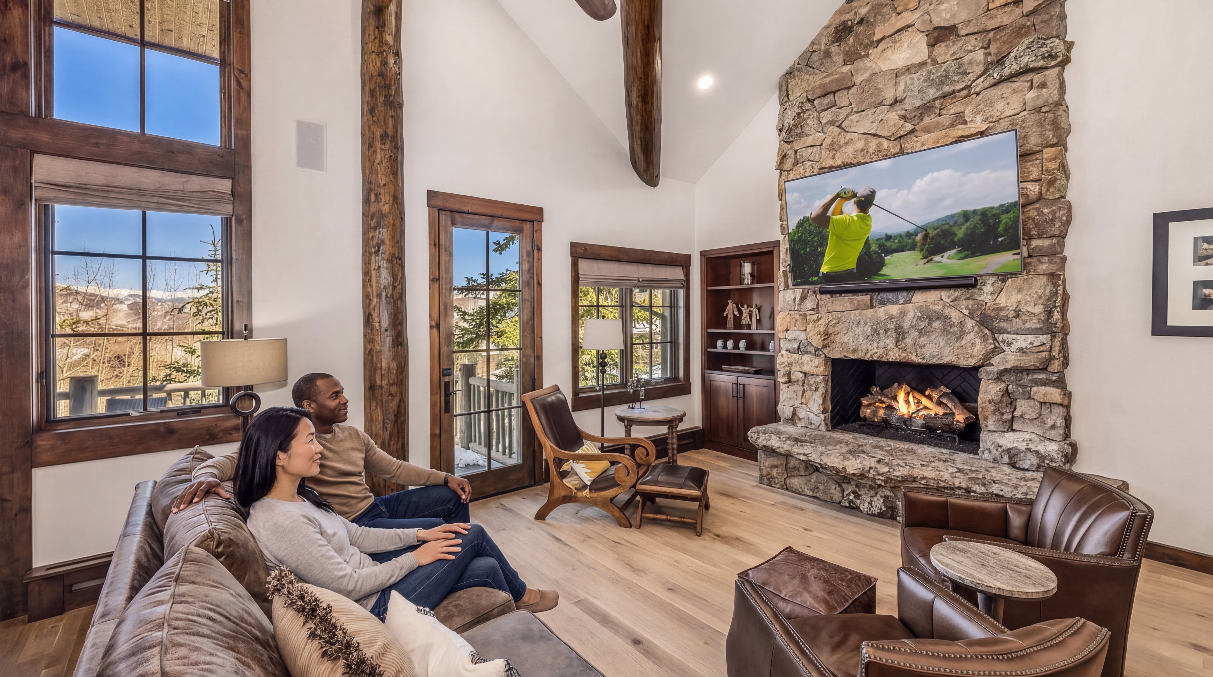 This cozy and inviting living room features a striking floor-to-ceiling stone fireplace as its centerpiece, topped with a wall-mounted television. The room is characterized by mountain-inspired rustic aesthetics, including exposed wood beams, arched windows with scenic views, and high-quality leather and wood furnishings. The open atmosphere and warm lighting create a luxurious yet comfortable space perfect for relaxation.