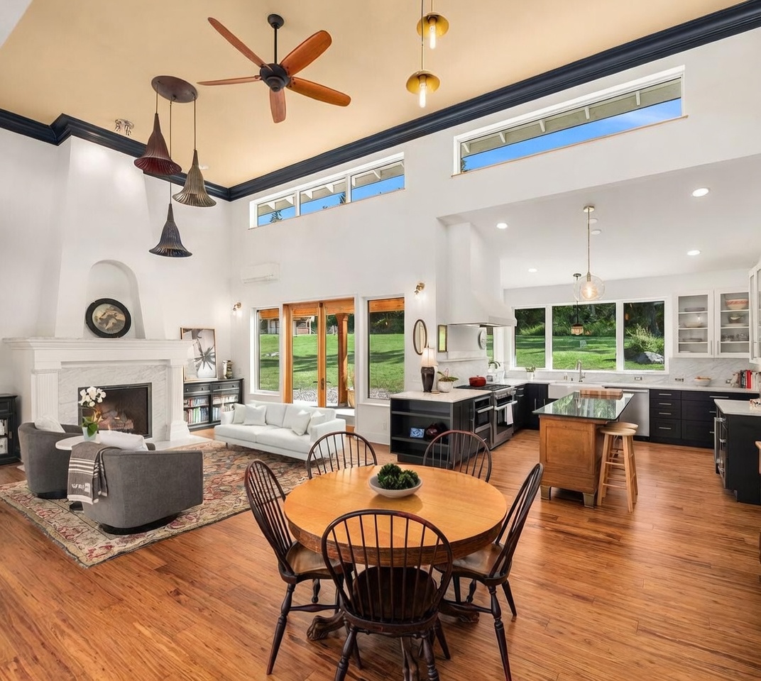 This open-concept living space features a high, vaulted ceiling with a central fan and a prominent fireplace with a white mantel and stone surround. The room flows seamlessly into a dining area with a round wooden table and a modern kitchen equipped with dark cabinetry and a central island. Large windows and glass doors provide abundant natural light and views of the lush green exterior, creating an airy and inviting atmosphere.