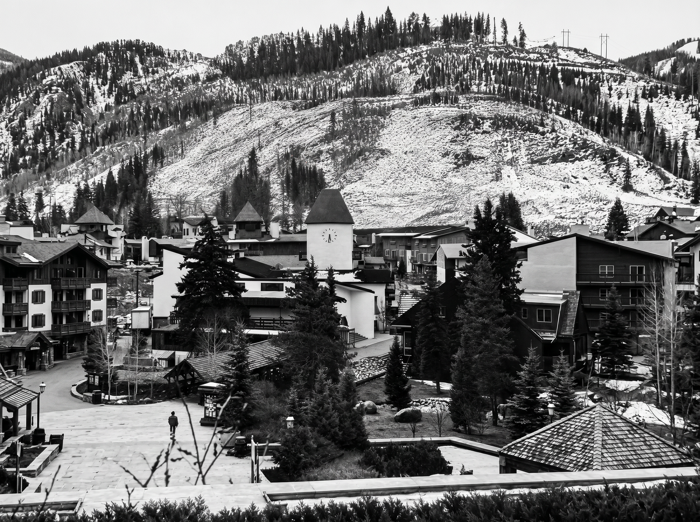 This black and white image captures a charming village-style community setting nestled at the base of a snow-dusted, tree-covered mountain. The center features a iconic clock tower building surrounded by interconnected paths, evergreen landscaping, and multi-unit residential structures. The aesthetic evokes a tranquil, resort-like atmosphere ideal for a mountain-side destination.