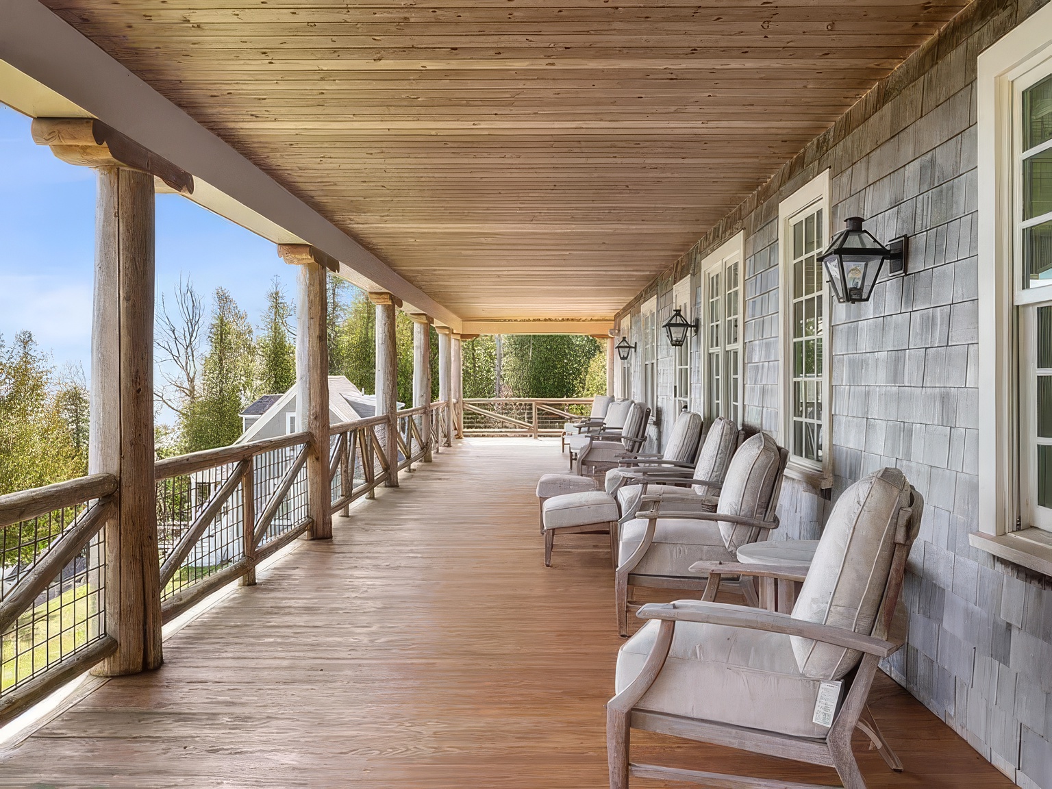 This expansive covered porch features a rustic aesthetic with natural wood support columns and a warm-toned wood plank ceiling. A row of comfortable outdoor armchairs with light-colored cushions is arranged along the gray shingled exterior wall, which is accented by classic wall-mounted lanterns. The deck overlooks a scenic, tree-lined landscape, providing a serene and inviting space for relaxation.