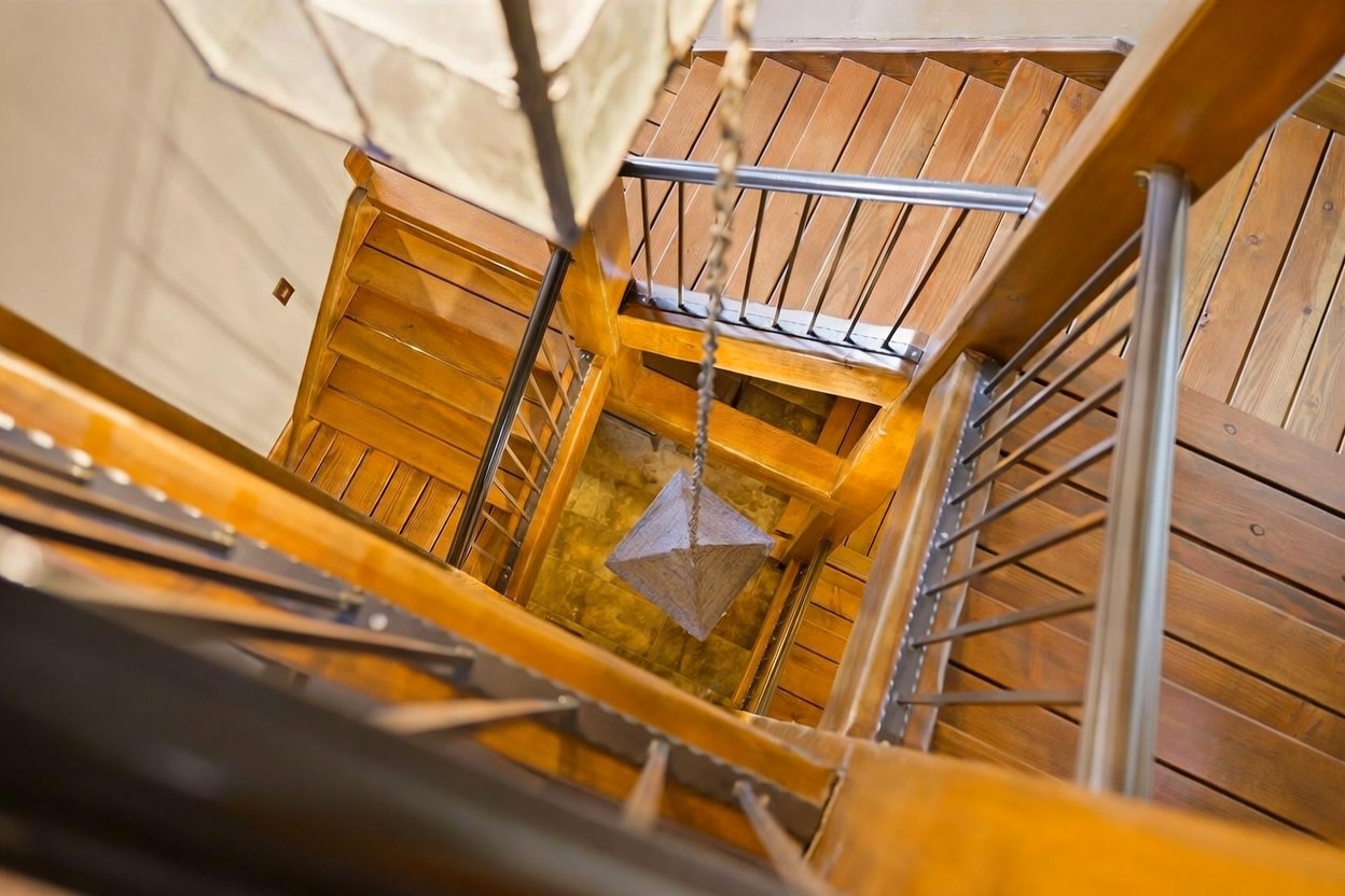 This high-angle, downward-looking perspective captures a striking wooden staircase winding around a central open void. The design features warm-toned wood treads and metal railings, creating a modern yet rustic aesthetic. A geometric pendant light hangs centrally within the stairwell, drawing the eye down toward the tiled landing below.