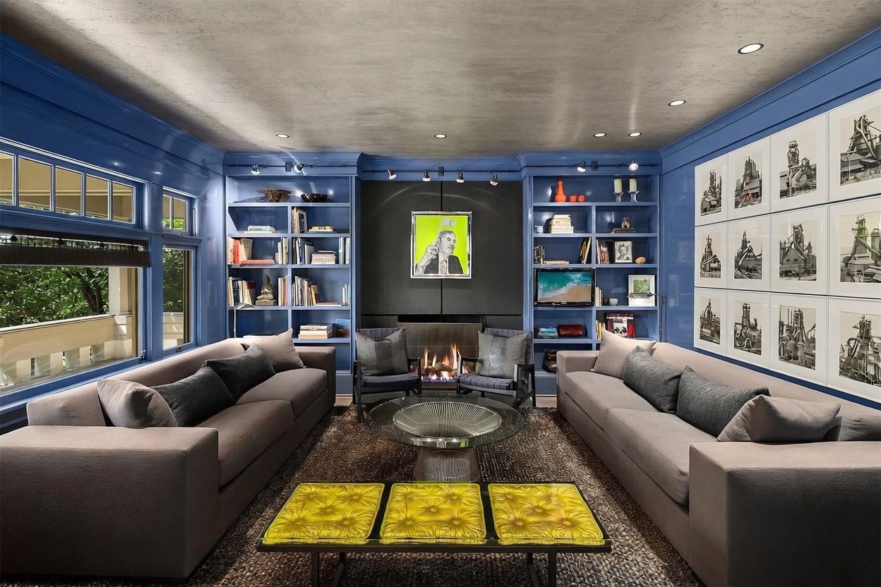 This sophisticated living room features bold blue built-in shelving flanking a central fireplace with a dark, textured surround and a vibrant piece of art. The space is anchored by two large, neutral-toned sofas facing each other, centered around a circular glass coffee table and a set of three yellow, textured ottomans. The room is illuminated by track lighting and a unique, mottled-finish ceiling, creating a moody and curated atmosphere.