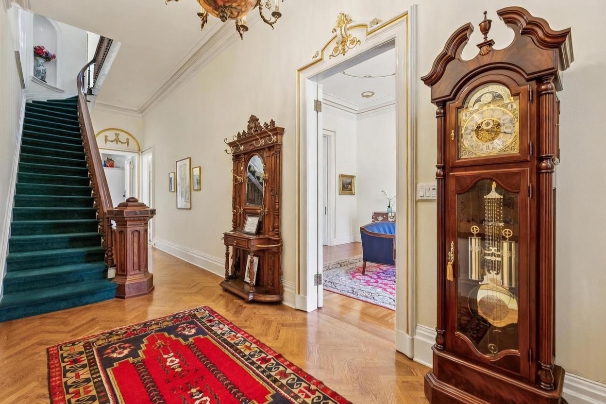 This elegant grand foyer features rich hardwood flooring, a sweeping staircase with a deep green runner, and a prominent grandfather clock. The space is adorned with ornate architectural details, including gold-trimmed door frames and a classic chandelier, creating a sophisticated and traditional atmosphere. A view through the doorway reveals a glimpse into an adjacent room with a patterned rug and blue seating, emphasizing the home's spacious and well-appointed layout.