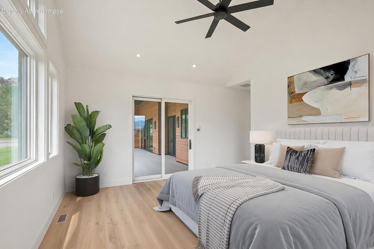 This bright and airy primary bedroom features light wood flooring and a neutral color palette, creating a serene atmosphere. A large bed with a textured headboard is positioned against a wall adorned with abstract art, while a tall potted plant adds a touch of greenery near the large windows. Sliding glass doors provide direct access to an outdoor deck, seamlessly blending indoor and outdoor living spaces.
