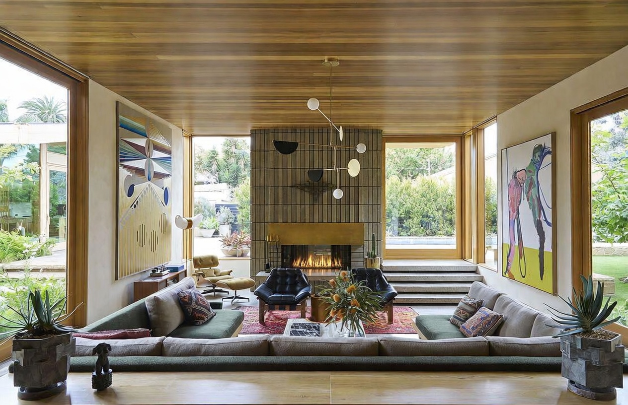 This sophisticated living room features a sunken seating area with a large, plush sectional sofa and two leather armchairs arranged around a central fireplace. The room is defined by its warm wood-paneled ceiling and expansive glass walls that provide views of the surrounding greenery. A striking modern chandelier hangs above the fireplace, which is framed by a textured, full-height stone surround, creating a focal point that blends mid-century modern aesthetics with contemporary luxury.