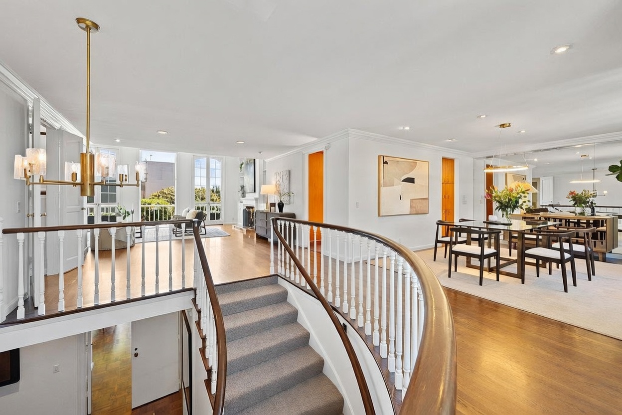 This bright, open-concept interior features a grand staircase with a curved wooden handrail and white balusters leading down to a lower level. The upper landing connects a formal dining area on the right, furnished with a modern table and chairs, to a spacious living area on the left that opens onto a balcony. The space is characterized by light wood flooring, neutral walls, and a large, elegant chandelier that serves as a focal point above the stairwell.