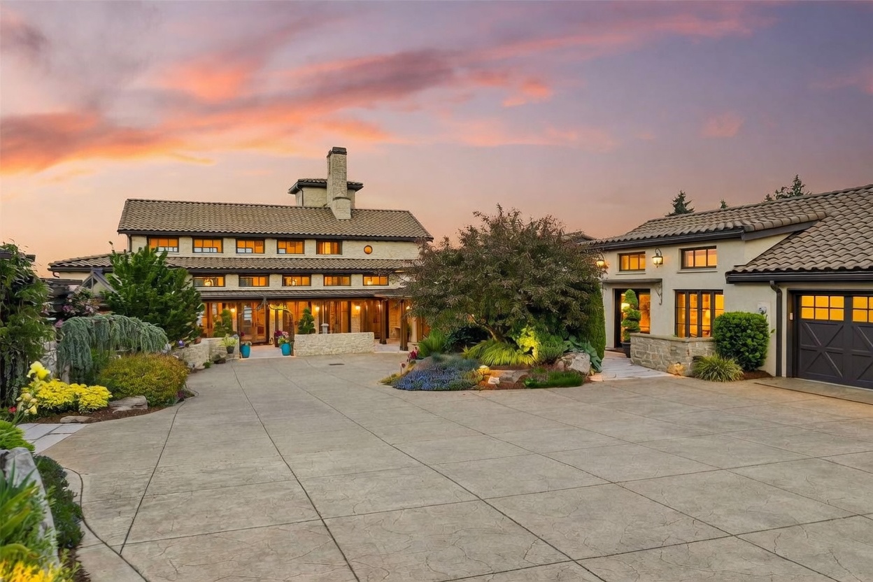 This expansive estate features a grand, multi-level residence with a Mediterranean-inspired aesthetic, characterized by light-colored stucco walls, stone accents, and a tiled roof. A large, paved motor court leads to the main entrance, framed by lush landscaping, mature trees, and decorative stone retaining walls. The scene is captured at dusk, with the warm glow of exterior lighting contrasting against a vibrant, colorful sunset sky.