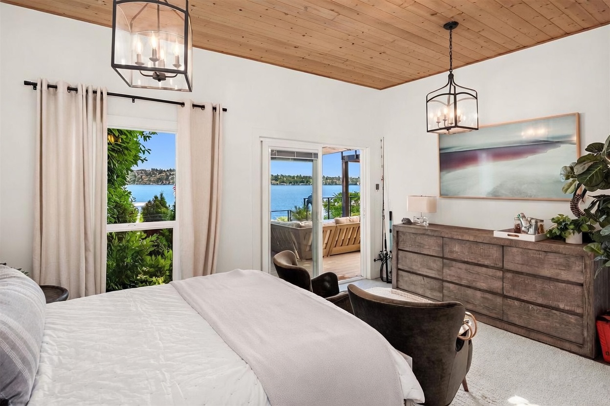 This primary bedroom features a serene, light-filled aesthetic with a wood-paneled ceiling and large windows that frame a scenic lake view. The room is furnished with a comfortable bed, two dark armchairs, and a substantial wooden dresser topped with a large, abstract landscape painting. Sliding glass doors provide direct access to an outdoor deck, seamlessly blending indoor and outdoor living spaces.