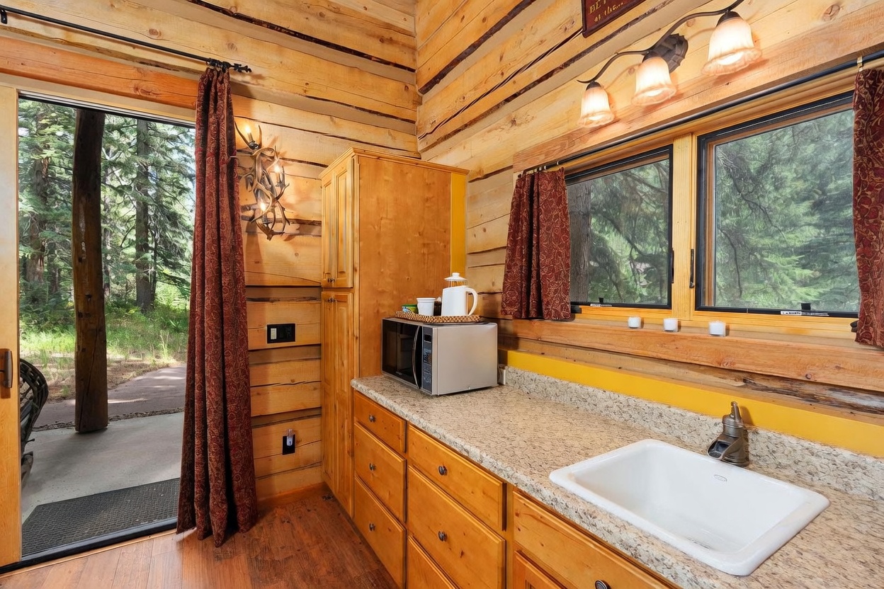 This rustic kitchen features light wood cabinetry, a speckled stone countertop, and a white farmhouse-style sink. The space is framed by log-style walls and includes a microwave, a coffee station, and a doorway leading directly to an outdoor forest view. Warm lighting from wall-mounted fixtures enhances the cozy, cabin-like atmosphere of the room.