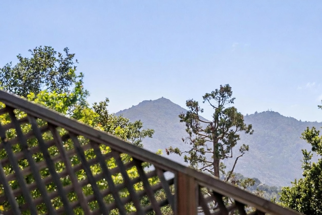 This perspective captures a scenic mountain view from a wooden deck or balcony, framed by a decorative lattice railing in the foreground. The composition highlights the natural beauty of the surrounding landscape, featuring lush green trees and distant, rugged peaks under a clear blue sky. The view emphasizes the property's elevated position and connection to the outdoors.