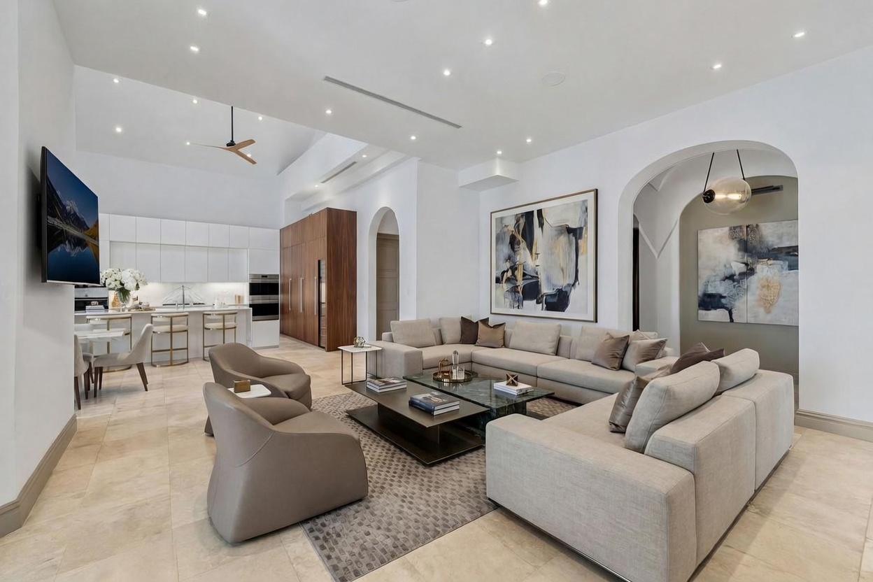 This expansive, open-concept living area features a neutral color palette with a large sectional sofa and two modern armchairs arranged around a central coffee table. The space flows seamlessly into a bright kitchen with white cabinetry and a breakfast bar, all under a high, vaulted ceiling. Large abstract artwork adorns the walls, and the room is illuminated by recessed lighting and a contemporary pendant fixture in the adjacent arched alcove.