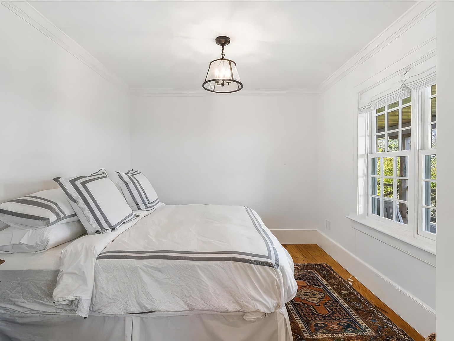 This bright and airy bedroom features crisp white walls and warm-toned hardwood flooring, creating a clean and inviting atmosphere. A plush bed with white bedding and striped accent pillows serves as the focal point, positioned beneath a classic pendant light fixture. To the right, a multi-pane window allows natural light to fill the room, while a patterned area rug adds a touch of texture and color to the space.